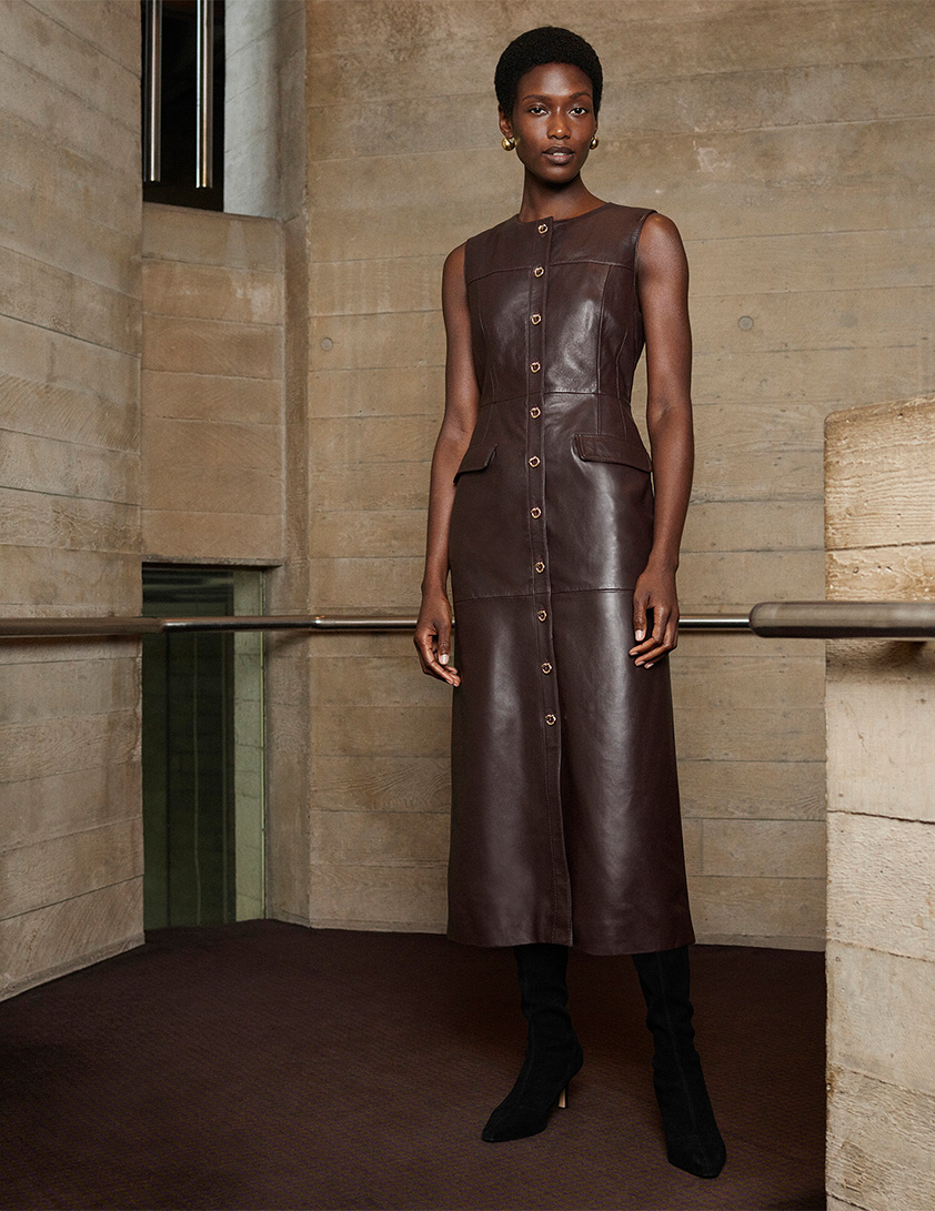 Woman wearing Whistles’ sleeveless chocolate-brown leather midi dress with button-through front and flap pockets, styled with gold earrings and black knee boots.