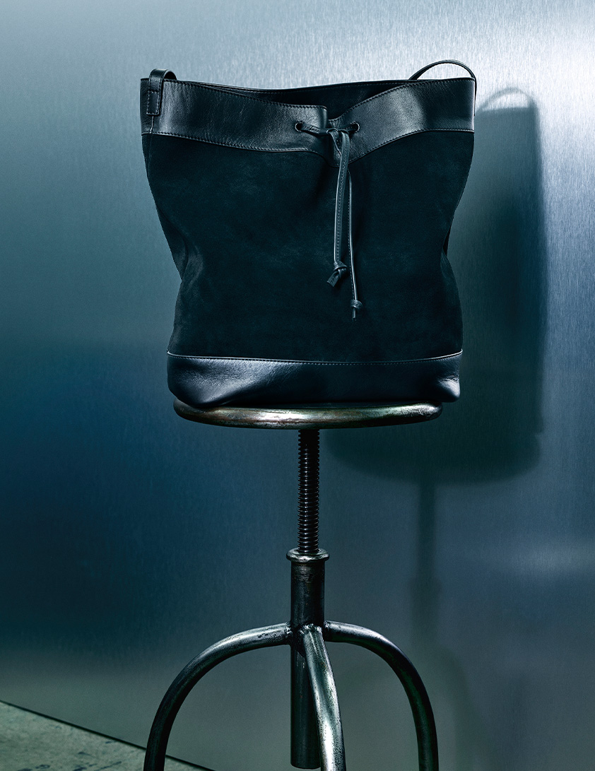 Whistles black suede and leather bucket bag with drawstring detail, displayed on a metal stool against a blue background.
