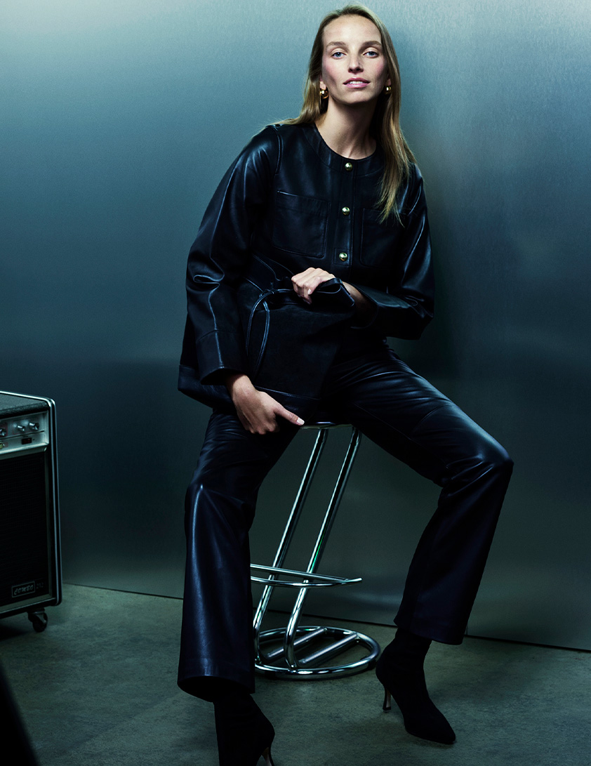 Woman wearing a Whistles black leather jacket with gold button detailing and matching pants, holding a black leather handbag