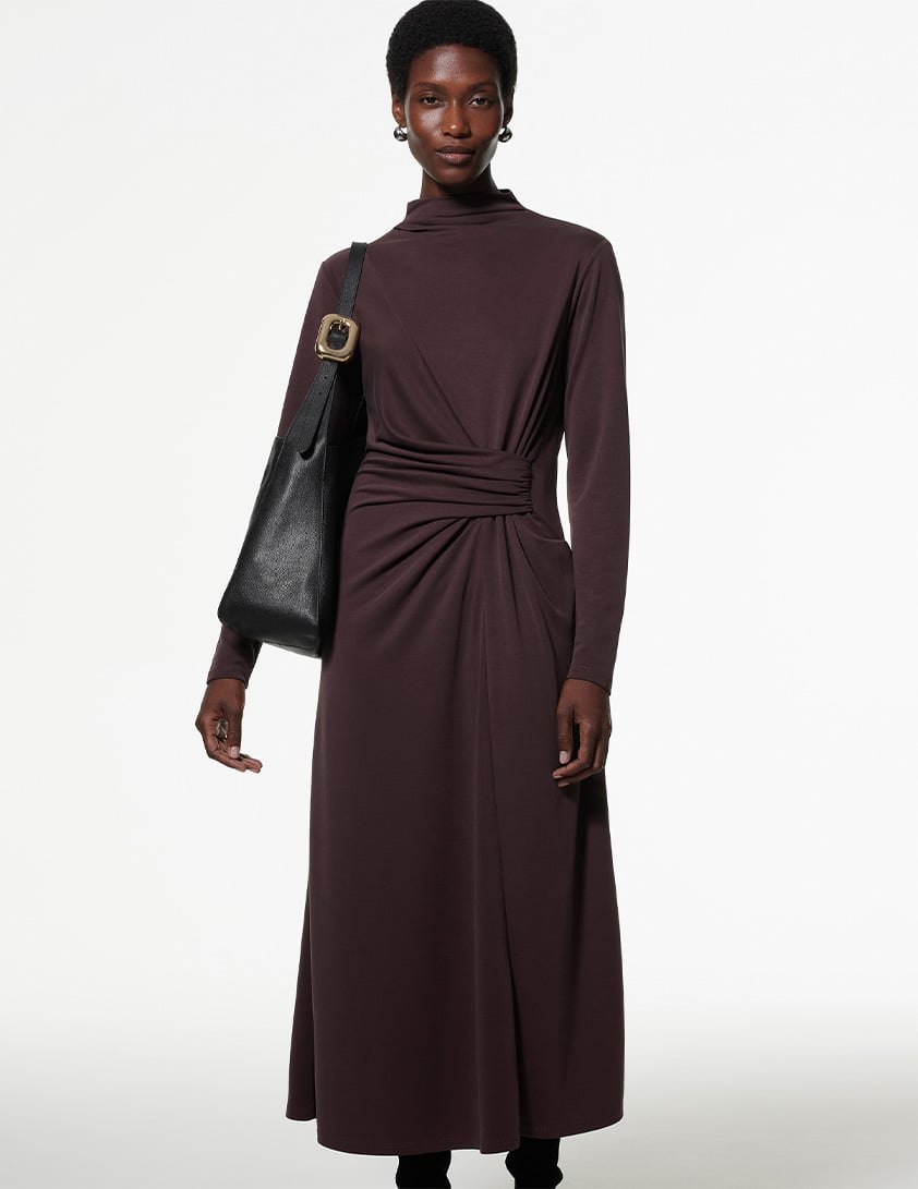 Woman wearing Whistles' chocolate-brown maxi dress with a twist-ruched waist, long sleeves, and a black leather shoulder bag