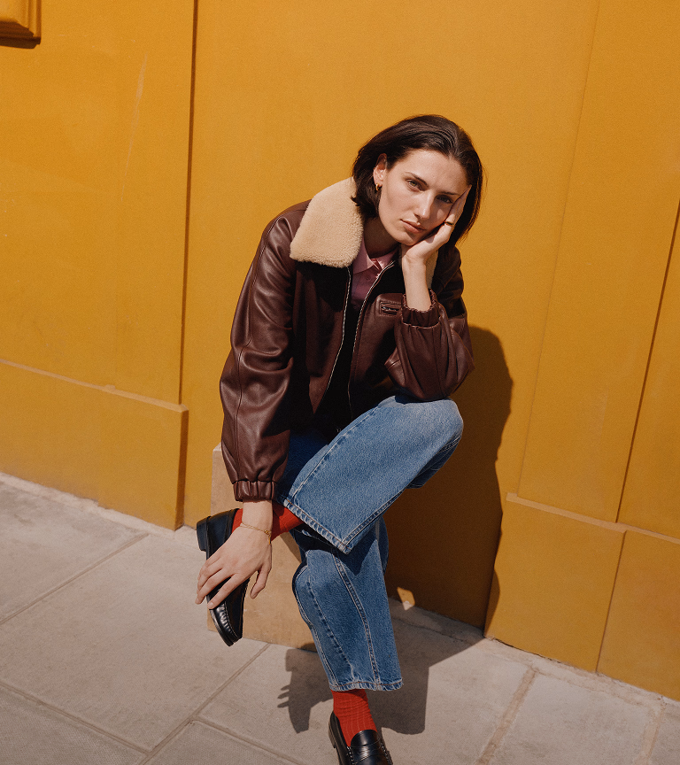 Model crouching against a yellow shopfront in a burgundy shearling-trim leather bomber and jeans