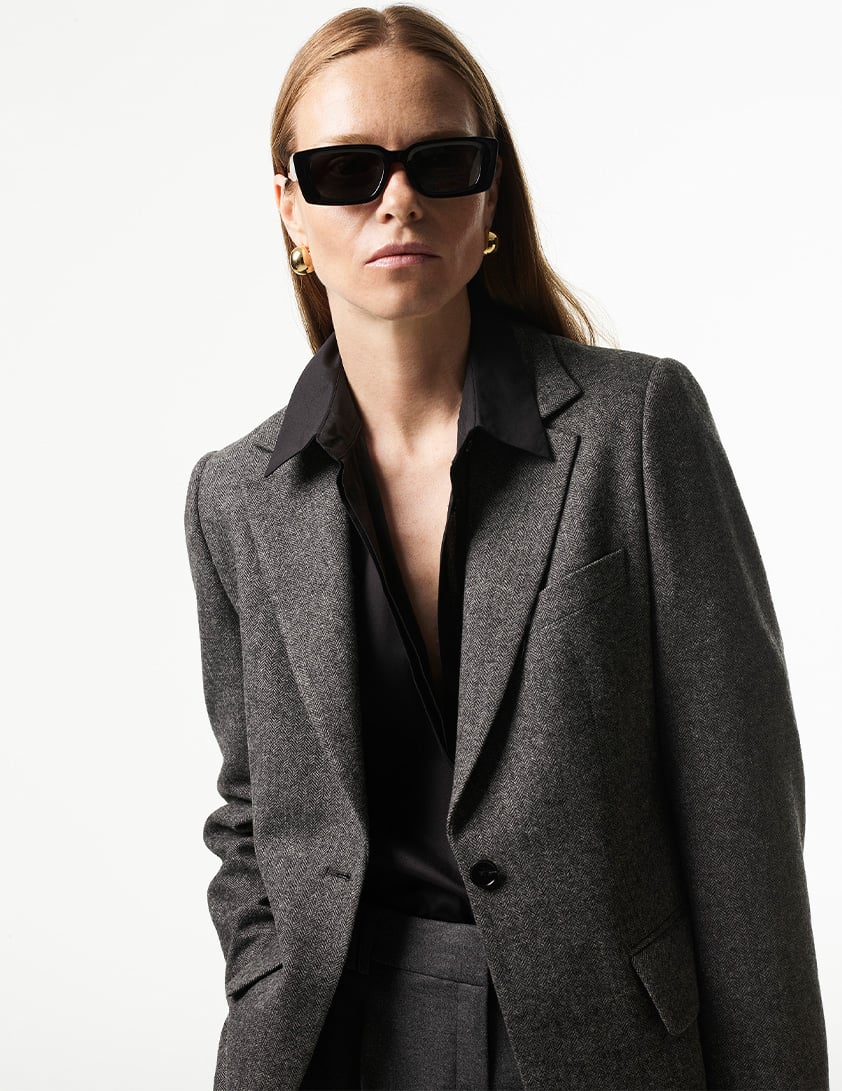 Woman wearing Whistles tailored grey blazer with black shirt gold hoop earrings and black sunglasses.