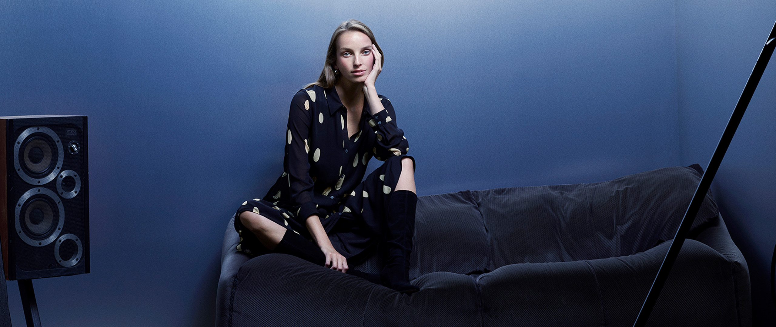 Woman sitting with arms crossed on a navy velvet sofa wearing a Whistles black polka dot dress