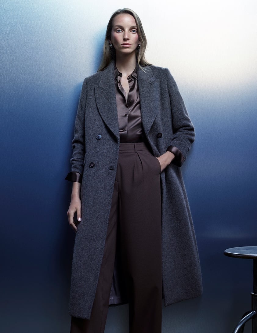 Woman wearing a Whistles grey double-breasted long coat with a brown satin shirt tucked into high waisted tailored pants