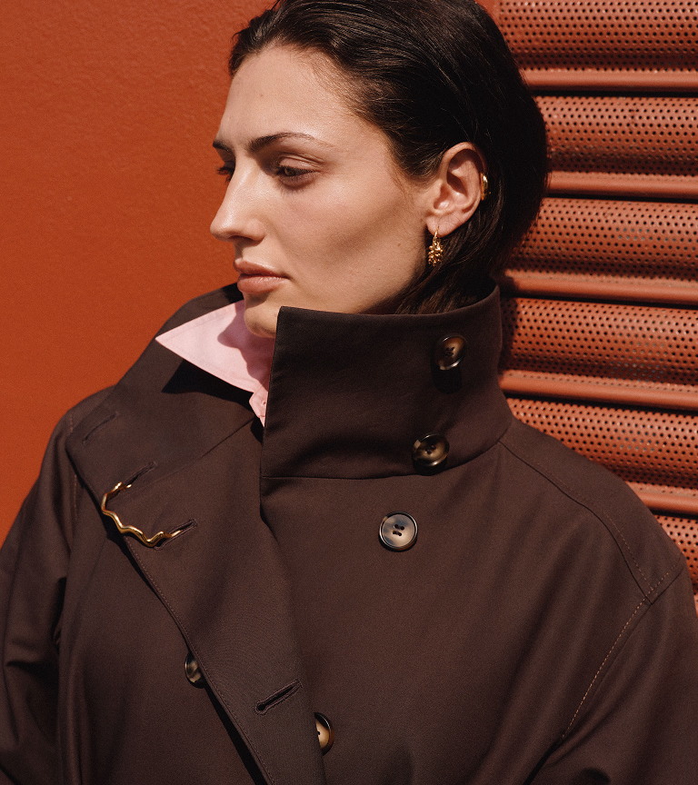 Model in a brown funnel-neck coat against a red mesh shutter