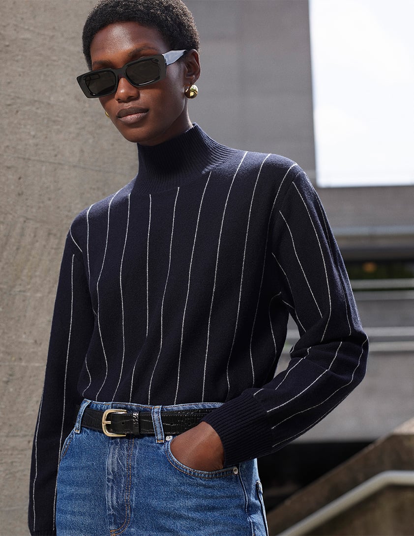 Woman wearing Whistles’ navy striped roll-neck knit sweater with mid-wash blue jeans, black belt, black sunglasses and gold earrings, part of Whistles’ 20% off sweaters promotion.