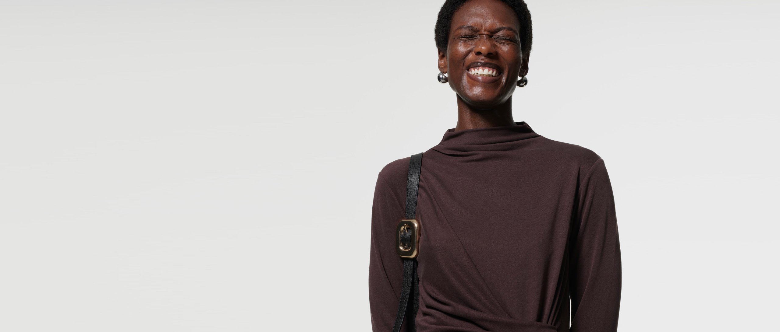 Woman wearing Whistles’ chocolate brown maxi dress with twist ruched waist and long sleeves styled with a black leather shoulder bag.