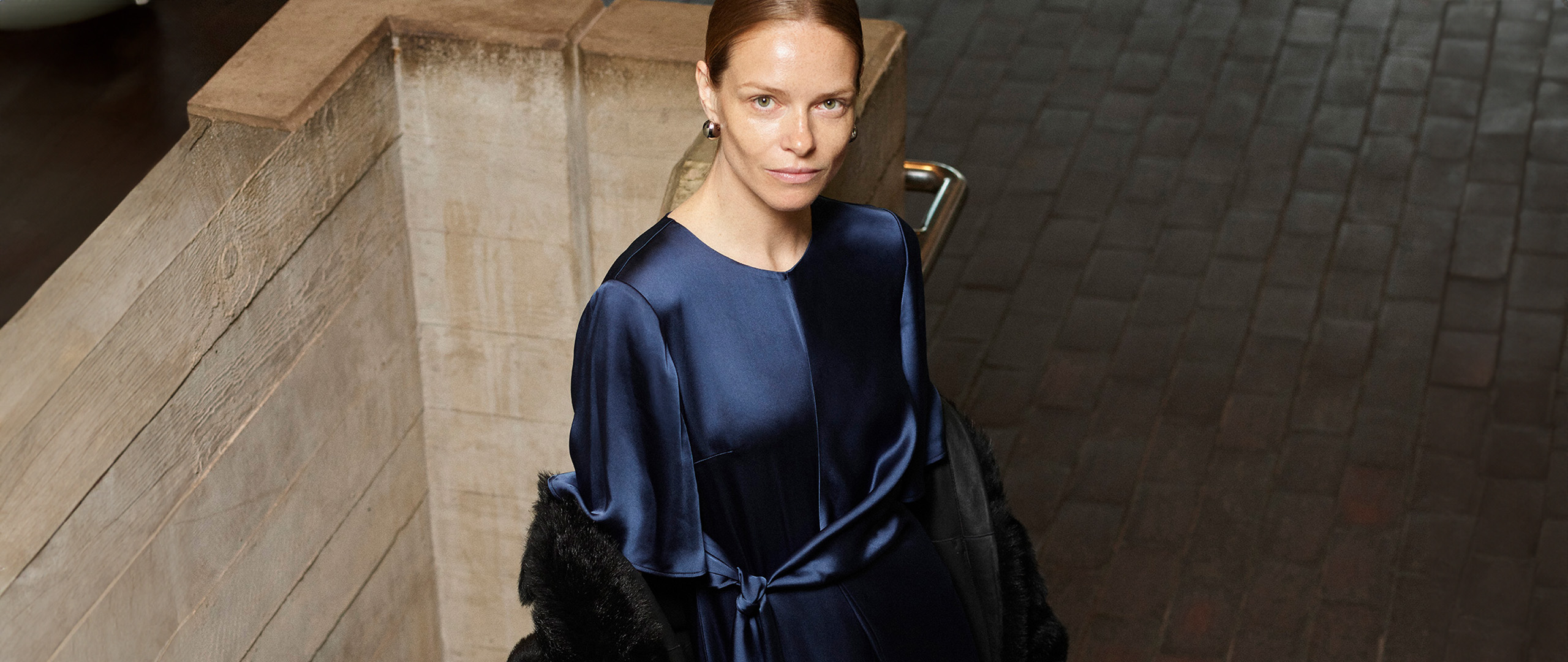 Woman wearing Whistles' satin navy midi dress with waist tie belt, styled with black faux fur coat, silver earrings and black boots