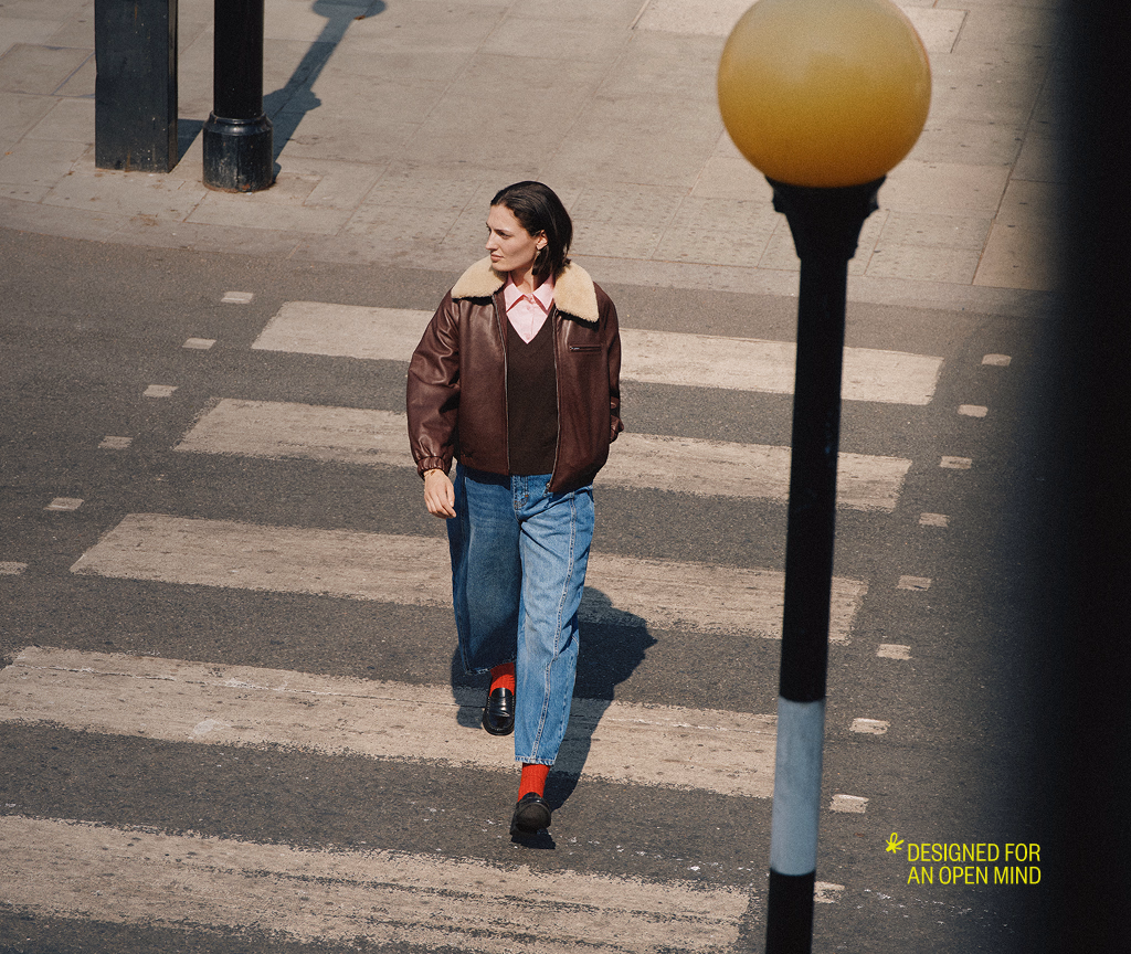 Model crossing a sunlit street past a lamp post, with the Designed For An Open Mind lockup