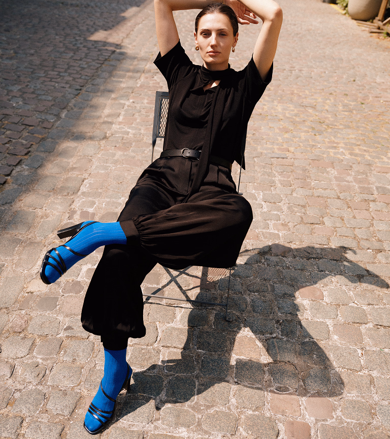 Emma sitting on a cobbled street in black satin balloon-leg trousers and blue heeled shoes